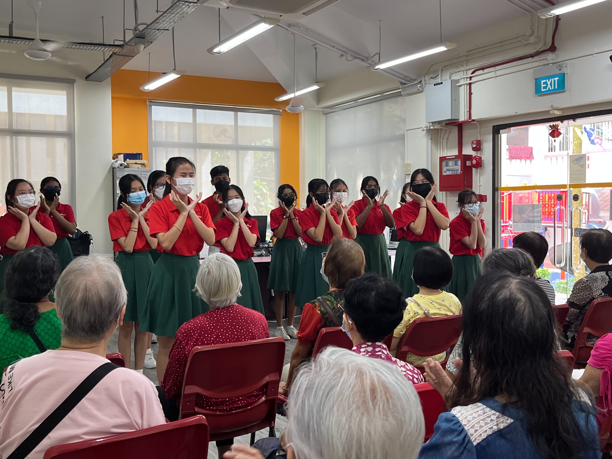 Students in red tops and green skirts wearing masks perform for seated elderly audience in an indoor community hall.